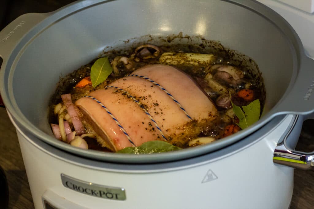 A small uncooked gammon joint resting in a slow cooker insert with sliced onion and a halved apple around it