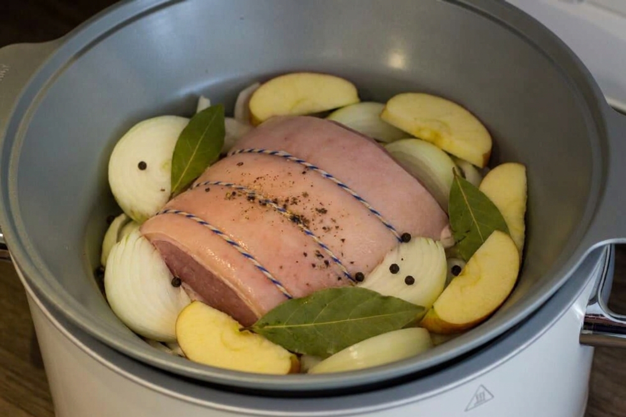 An uncooked gammon joint in a slow cooker surrounded by thick onion slices and apple wedges before cooking
