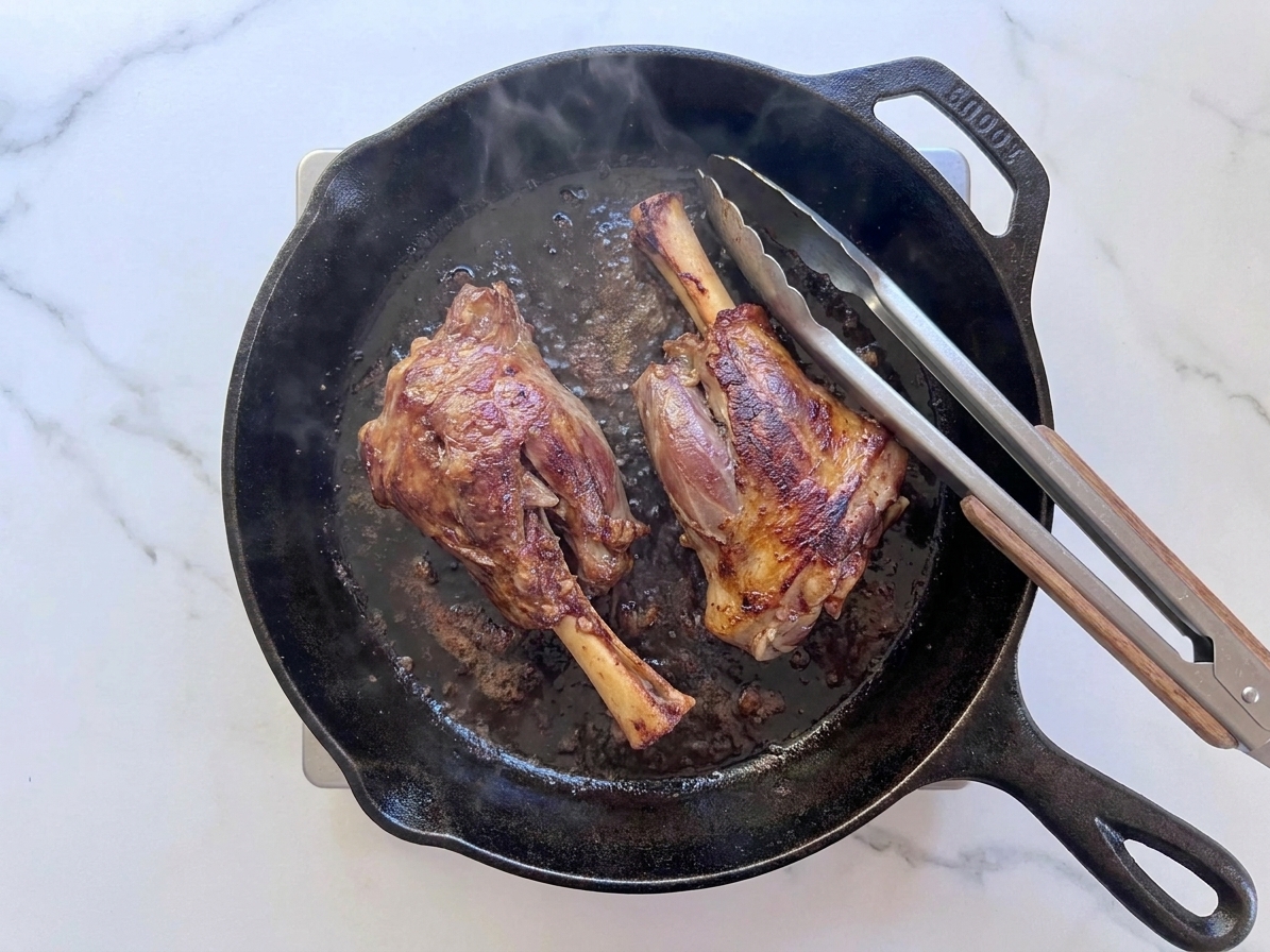 Two lamb shanks browning in a cast iron skillet with visible caramelized edges and a pair of tongs resting on the pan handle